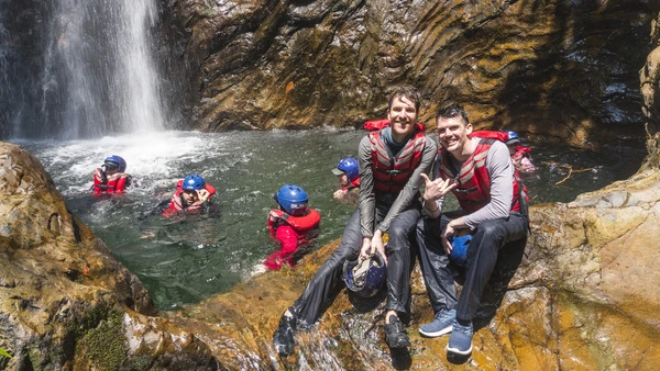 Two happy men by the waterfall with other people swimming in the cascade.